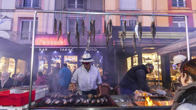 Homme faisant griller des harengs devant le bar l'Escale sur le quai Henri 4. Des harengs crus sont accrochés à une barre au-dessus du grill. A l'arrière-plan des personnes sont attablées en terrasse