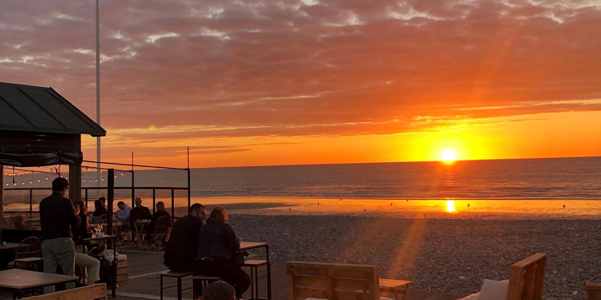 Terrasse d'un bar sur les galets devant un coucher de soleil face à la mer