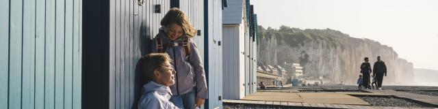 Une maman et sa fille sont assises devant une cabine de plage bleue à Dieppe et rient ensemble, avec les falaises et des promeneurs en arrière-plan