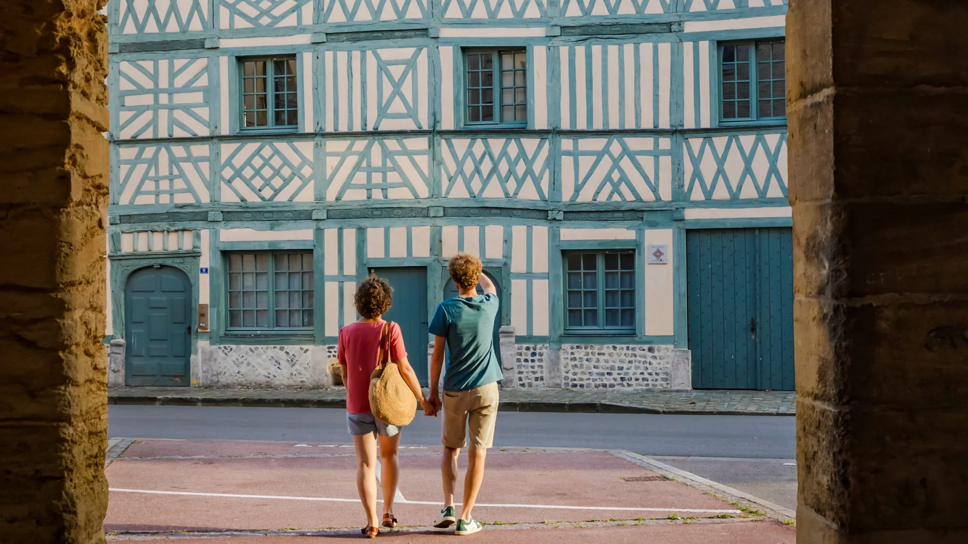 Deux visiteurs découvrent la Maison Miffant à Dieppe, remarquable façade à pans de bois, vue sous une arche en pierre.