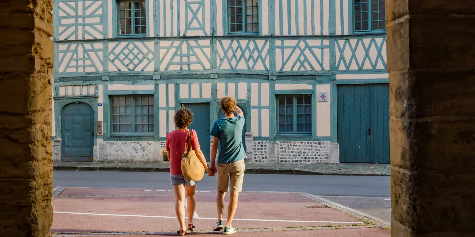 Deux visiteurs découvrent la Maison Miffant à Dieppe, remarquable façade à pans de bois, vue sous une arche en pierre.