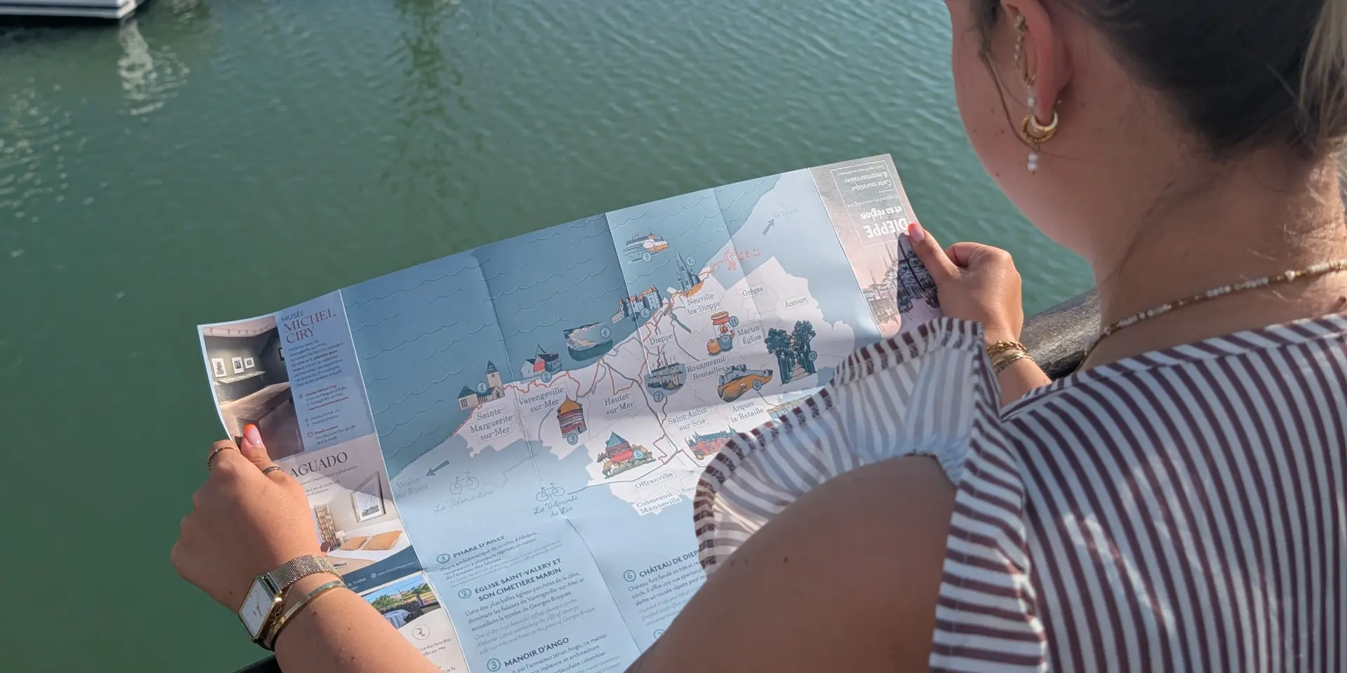 Visiteuse consultant une carte touristique illustrée face au port de plaisance de Dieppe, avec les voiliers et les façades du quai Henri IV en arrière-plan.