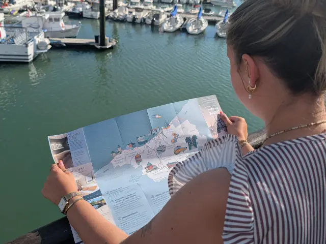 Visiteuse consultant une carte touristique illustrée face au port de plaisance de Dieppe, avec les voiliers et les façades du quai Henri IV en arrière-plan.