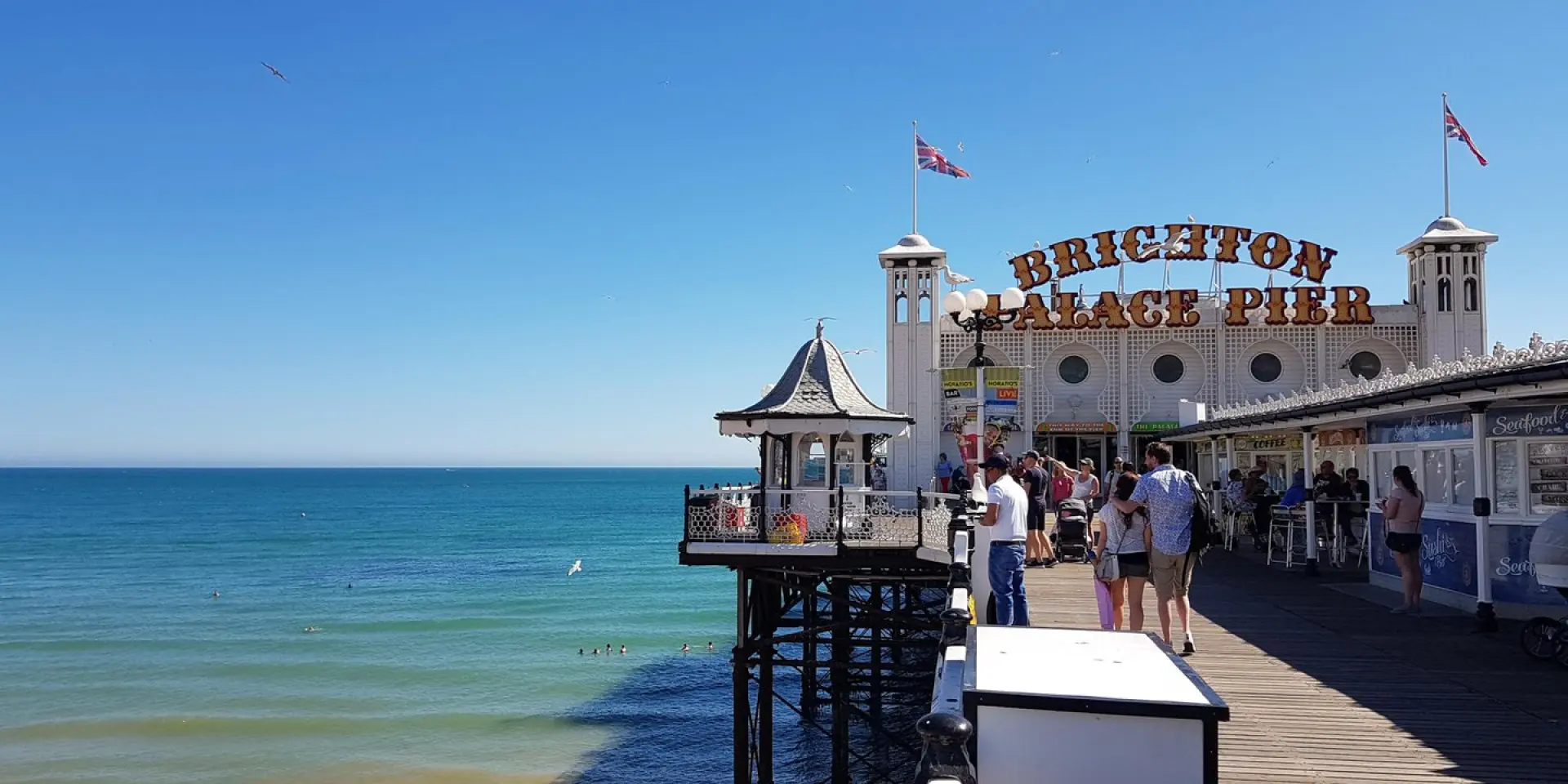 Visiteurs se promenant sur le Brighton Palace Pier, au-dessus de la plage et de la mer, sous un ciel bleu parcouru de mouettes.