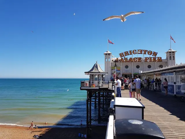 Visiteurs se promenant sur le Brighton Palace Pier, au-dessus de la plage et de la mer, sous un ciel bleu parcouru de mouettes.