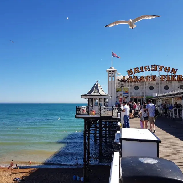Visiteurs se promenant sur le Brighton Palace Pier, au-dessus de la plage et de la mer, sous un ciel bleu parcouru de mouettes.