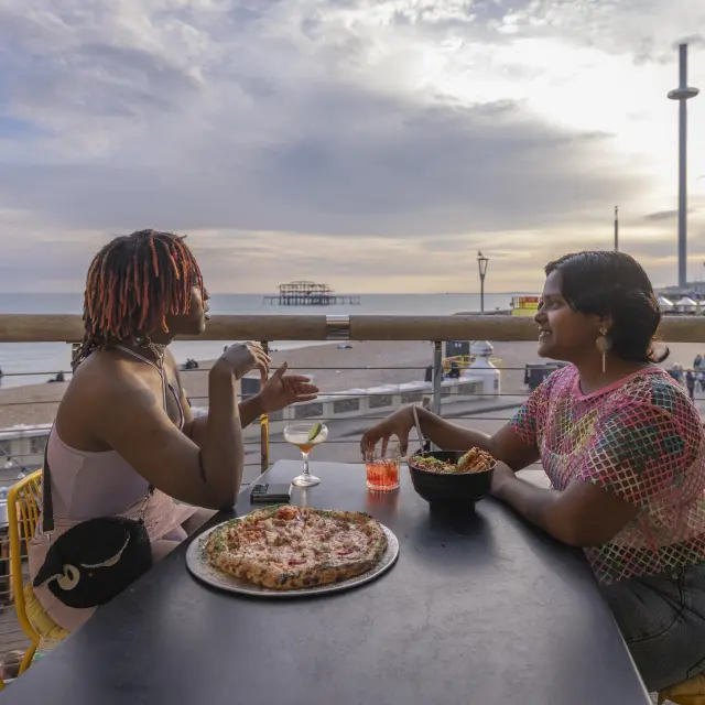 Deux visiteuses partagent un repas en terrasse au Shelter Hall à Brighton, avec la plage, la mer et le West Pier en arrière-plan