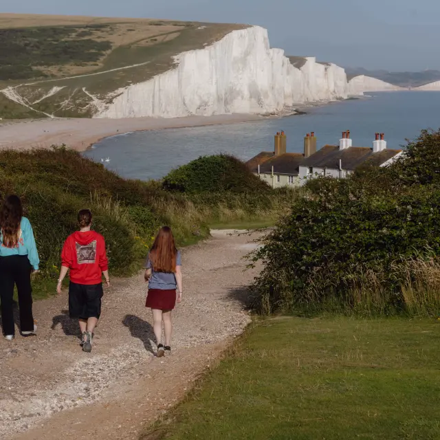 Randonneurs descendant un sentier vers la côte, avec les falaises blanches des Seven Sisters dominant la mer en arrière-plan.A selection of photographs taken at Buzz Active, Towner Gallery and Birling Gap in East Sussex. A series of images to promote travel experiences in East Sussex. PHOTOGRAPHY BY DIENSEN PAMBEN.