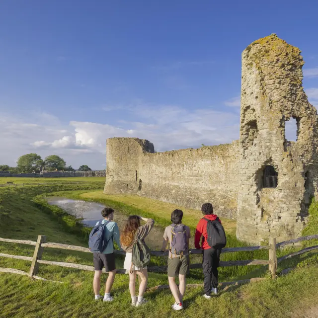 Quatre visiteurs découvrent les ruines de Pevensey Castle, entourées de douves herbeuses sous un ciel bleu.